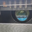 Detail kytarového komba s viditelným reproduktorem a částí elektroniky za mřížkou. Na středovém krytu reproduktoru je bílé logo Peavey a modrý nápis…