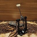 Fotografie zobrazuje černý basový pedál s kovovými mechanismy a řetězem. Na šlapce jsou vyfrézované otvory a nečitelné logo. Beater je zakončen bílou…