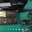 Černý digitální metronom a ladička s displejem, na kterém je nápis musedo MT-80 METRONOME & TUNER. Zařízení má několik ovládacích tlačítek s popisky…