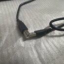 Černý USB kabel s konektory typu A a B. Na plastovém krytu konektoru je vyryto logo USB. Kabel vykazuje běžné známky používání, povrch plastu je matn…
