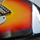 Detail korpusu elektrické kytary s barevným přechodem od žluté přes červenou k černé. Viditelný je kovový most sádlů a část bílého pickguardu. Povrch…