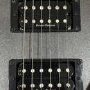 Detailní pohled na dva elektrické snímače typu humbucker v černých rámečcích, osazené na stříbrném kovovém povrchu s texturou. Oba snímače mají černé…