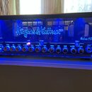 Kytarový zesilovač s černým rámem a modrým podsvícením, uvnitř kterého jsou vidět elektronické lampy. Na předním skle v centru je bílý nápis Hughes &…