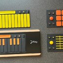Fotografie zobrazuje čtyři moduly elektronického hudebního kontroléru s černým základem a barevnými prvky. Jeden modul má žluté klávesy, druhý oranžo…