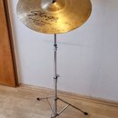 Zlatý činel umístěný na chromovaném stojanu. Na povrchu jsou nápisy „Cx“, „Stagg“, „Hand Made“ a „18" Crash-Ride CXCR-18“. Povrch činelu vykazuje dro…