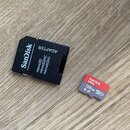 Černý adaptér s nápisy SanDisk, ADAPTER a microSD microSDHC microSDXC. Vedle něj je malá paměťová karta s červeným pruhem a nápisy SanDisk Ultra, 128…