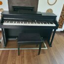 Černý digitální piano v černém dřevěném korpusu s přívodní lavičkou s černé polstrováním. Nad klávesnicí je umístěno ovládací panel s tlačítky a disp…
