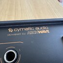 Detailní záběr na černý povrch elektronického zařízení. Je na něm vidět bílý nápis „cymatic audio“ s doprovodným logem květiny a pod ním text „powere…