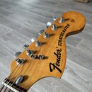 Fotografie zachycuje hlavu elektrické kytary z přírodního dřeva s lakovaným povrchem. Na hlavě je černý nápis Fender® STRATOCASTER a menší text PAT.…