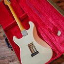 Bílá elektrická kytara s tvarem typu Stratocaster v červeném plišovém kufru. Nástroj má světlý dřevěný krk a hmatník, na zadní straně těla je vidět k…