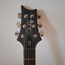 Fotografie zachycuje hlavu černé elektrické kytary s chromovanými ladičkami. Na povrchu je bílý nápis "Paul Reed Smith SE" a vertikálně orientovaný n…