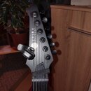 Fotografie zachycuje hlavu černé elektrické kytary s nápisem SPIRA GUITARS. Na hlavě je upevněn černý kapestry s logem BOSS. Povrch nástroje má matný…