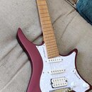 Bordeaux červená elektrická kytara s matným povrchem a bílým pickguardem. Na hmatníku je vidět nápis „strandberg“. Nástroj má tři bílé snímače, zlatý…