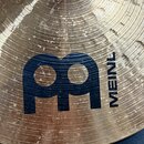 Detailní záběr na povrch mosazného činele s viditelným rakováním a koncentrickými drážkami. Na povrchu je umístěno černé logo ve tvaru stylizovaného…