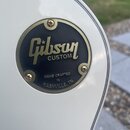 Detailní záběr na kulatý černý štítek s kovovým zlatým rámem, připevněný na bílý povrch nástroje čtyřmi šrouby. Na štítku je zlatým písmem nápis Gibs…