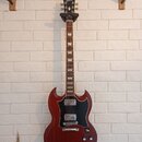 Elektrická kytara s červeným trupem v charakterickém tvaru SG a černým pickguardem. Na hlavě hmatníku je bílý nápis Gibson. Nástroj má dva humbuckery…
