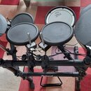 Elektronická bicí souprava černé barvy na kovovém stojanu. Na padech a spodní hraně stojanu je vidět bílý nápis Roland. Souprava obsahuje čtyři pady…