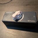 Černý efekt pedál s bílým ovládacím knoflíkem. Na horní ploše jsou nápisy VOLUME, OUT, IN a symbol +. V pravém dolním rohu je logo MH pedals. Povrch…