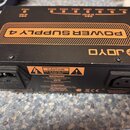 Černý napájecí zdroj s oranžovými prvky a nápisem POWER SUPPLY 4. Na horní straně je viditelné logo JOYO a řada výstupů s označením mA. Na zadní stra…