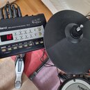 Fotografie zachycuje část elektronické bicí soupravy. Je zde vidět černý modul s nápisy Roland, PERCUSSION SOUND MODULE TD-3 a V-Drums, který má zapn…