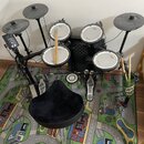 Elektronická bicí souprava značky Roland s černým rámem a gumovými pady. Na padesátích padech je vidět nápis Roland a V-Drums. Součástí je černý sedá…