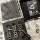 Tři černé elektrické snímače pro kytaru s mosaznými póly, umístěné v černé pěnové vložce. K snímačům vedou elektrické vodiče v různých barvách. Vedle…