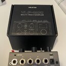 Fotografie zobrazuje stříbrný sampler s černými ovážnými knoflíky a konektory, umístěný před svým černým originálním krabicí. Na krabici je bílý nápi…