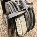 Černý kabel namotaný na plastovém návijáku s rukojetí. Na konci kabelu je velký vícepinový konektor v šedém kovovém krytu. Povrch plastu a kovového k…