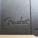 Detailní záběr na černý plastový povrch s texturou, pravděpodobně součást pouzdra nebo kufru. Uprostřed je vyražené logo Fender®. Povrch vykazuje dro…