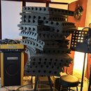 Stoh tmavě šedých akustických pěnových panelů s různými povrchovými strukturami (vlnovité a pyramidové). Vedle stohu stojí kytarový kombozesilovač s…