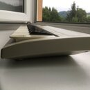 Bílá elektronická klávesová klaviatura s plastovým šasi. Nástroj je umístěn na parapetu u okna. Na viditelných částech nejsou žádné čitelné značky, l…