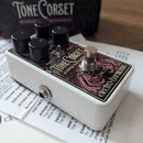 Bílý efekt pedál s nápisy Tone Corset a analog compressor. Na pravém boku je umístěno logo electro-harmonix. Zařízení má čtyři černé ovládací knoflík…