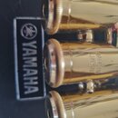 Detailní záběr na tři zlaté ventily trubky. Na černém podkladu je vidět logo a nápis YAMAHA. Na prostředním ventilu je vyražen text MADE IN JAPAN. Po…
