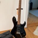 Černá elektrická kytara s hnědým hmatníkem a světlou hlavou, umístěná na stojanu. Nástroj má tři snímače typu humbucker a stříbrný hardware. Na spodn…