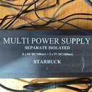 Černý obdélníkový napájecí adaptér s množstvím připojených černých kabelů. Na horní straně je bílý nápis MULTI POWER SUPPLY, SEPARATE ISOLATED a 8 x…