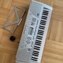 Světle šedý elektronický klavír s plastovým korpusem a bílo-černými klávesami. Na přístroji jsou viditelné nápisy „128 TONE“ a „128 VOICE“, uprostřed…
