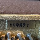 Detailní záběr na zlatý, strukturovaný povrch pouzdra s hnědým vnitřním polstřením. Uprostřed je vidět čitelný nápis 159821. Ve spodní části jsou vid…