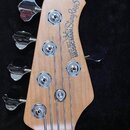Fotografie zachycuje hlavu pětistrunné basové kytary z přírodního dřeva s chromovanými ladičkami. Na povrchu je svisle napsán text ERNIE BALL MUSIC M…