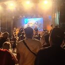 Fotografie zachycuje koncertní scénu s kapelou a publikem z pohledu diváka. Na pódiu jsou vidět hudebníci s nástroji, v pozadí velká LED obrazovka a…