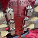 Zadní strana červené hlavy kytary s nápisem 221140457 a textem MADE IN USA. Na hlavě jsou instalovány šest kovových ladících kolíků s krémovými knofl…
