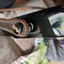 Detailní záběr na černý lakovaný povrch hudebního nástroje. K kovovému úchytu je připevněn hnědý kožený řemen s viditelným bílým stehem. Na povrchu n…