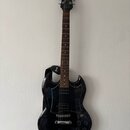 Černá elektrická kytara s tmavým hnedým hmatníkem a černým pickguardem. Na hlavě nástroje je bílý nápis Epiphone. Kytara má připevněný černý popruh s…