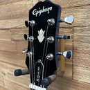 Černá hlava elektrické kytary s bílým nápisem Epiphone a stříbrným logem ve tvaru stylizovaného listu. Na hraně hlavy je svisle napsáno SG MUSE. Nást…
