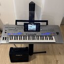Šedý digitální klavír s logem YAMAHA na horní části a označením Genos na pravé straně. Nástroj je umístěn na černém stojanu a nad ním je průhledlá hu…