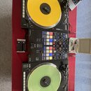 Sada pro DJing složená ze dvou černých gramofonů, mixážního pultu a příslušenství na červeném podkladu. Horní gramofon má žlutý vinyl, dolní světle z…