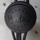 Kruhové černé plastové příslušenství s připevněnými černými šňůrkami. Na povrchu je vyražen nápis WWW.JAZZLAB.COM, logo jazzlab a slovo patented. Pov…
