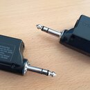 Dvě černé plastové bezdrátové vysílače s kovovými 6,3mm jack konektory. Na jednom je vidět nápis „TRANSMITTER WL-1 ONLY“ a „To AMPLIFIER“ s ikonou ze…