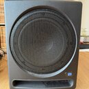 Černý subwoofer s hranatým korpusem a zaoblenými rohy. Přední strana je kryta velkou kulatou kovovou mřížkou s jemným perforovaným vzorem. V pravém d…