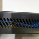 Detailní záběr na černý kovový šasi s ventilačními otvory ve tvaru šikmých rýh. Uvnitř zařízení jsou vidět modré kabely a části elektronických kompon…