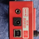 Detail červeného kovového zařízení s konektory. Na bílém štítku jsou nápisy AUX IN, DC IN USE BOSS PSA ADAPTOR ONLY a USB s doprovodným symbolem. Jso…